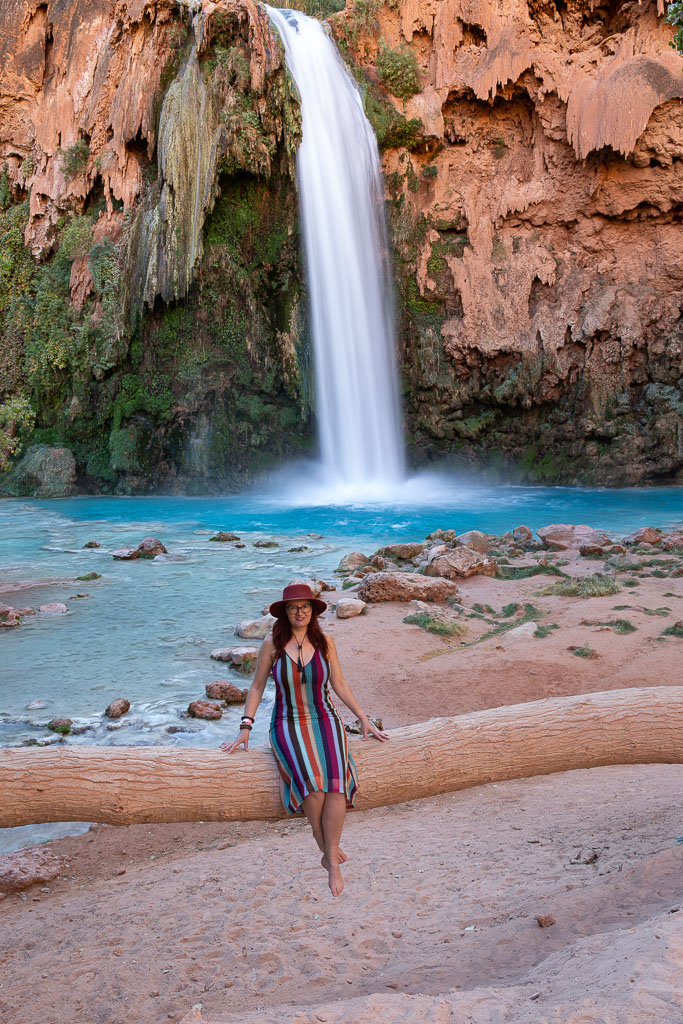 Agnes Stabinska, the author, sits in a colorful striped dress and a brown hat on the trunk of a large tree with the Havasu Falls waterfall in the background. A blue water flows down the orange wall.