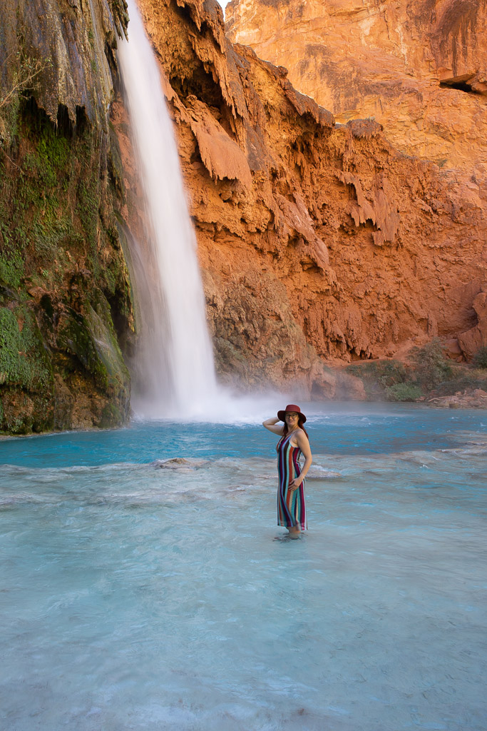 Agnes Stabinska, the author, stands calf-deep in the blue water of Havasu Falls. Large streams of water flow from the orange wall. Agnes has a brown hat and a striped dress.