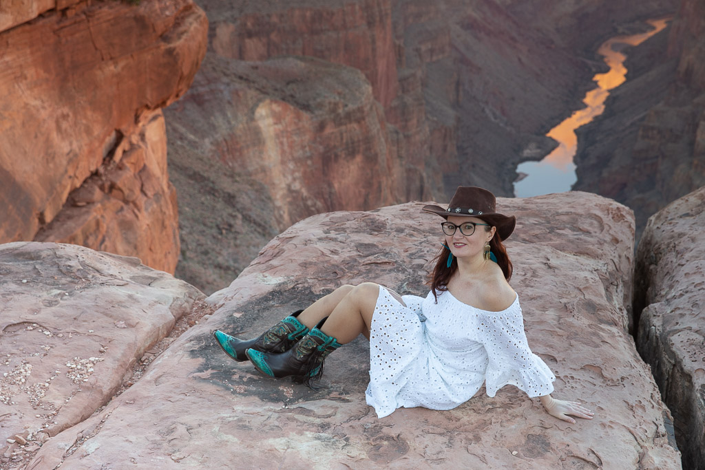 Toroweap Overlook—Agnes Stabinska, the author, in a white dress, brown cowboy boots, and brown cowboy hat, is sitting on the edge of the overlook and admiring the views: the Colorado River at the bottom and stunning canyon walls.