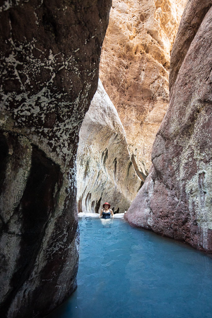 Agnes Stabinsa, the author, is immersed in the blue water of Aricona Natural Hot Springs in a brown hat and a black swimsuit. The pool with water is located between the high orange walls of the canyon