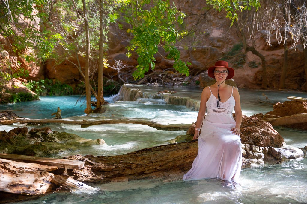 The author, Agnes Stabinska, in white dress and brown hat is siting on a tree in the water and relax in Beaver Falls at Havasupai.