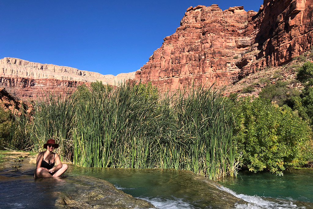 The author, Agnes Stabinska, sits in a black swimsuit and a brown hat, immersed in the blue water of the falls among orange rocks and greenery.