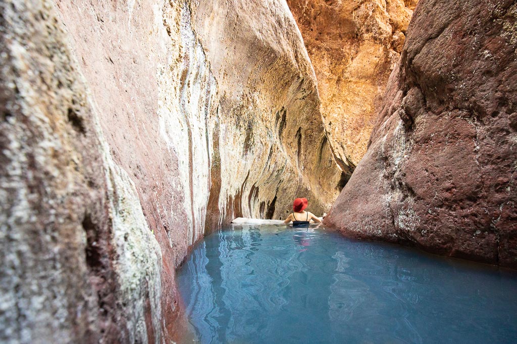 Agnes Stabinsa, the author, is immersed in the blue water of Aricona Natural Hot Springs in a brown hat and a black swimsuit. The pool with water is located between the high orange walls of the canyon