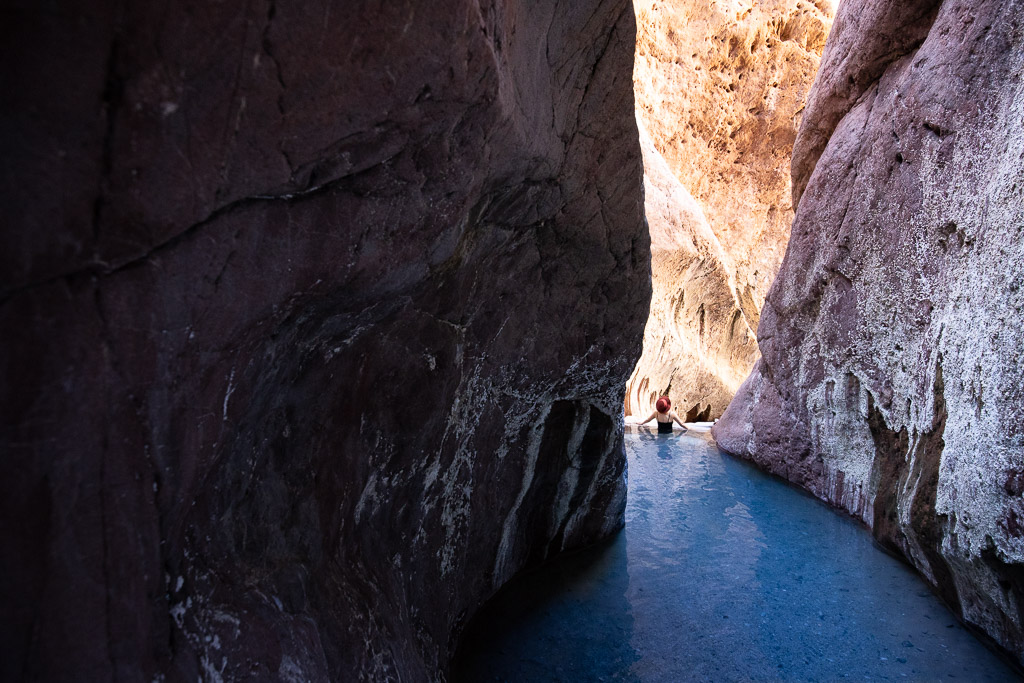 Agnes Stabinsa, the author, is immersed in the blue water of Aricona Natural Hot Springs in a brown hat and a black swimsuit. The pool with water is located between the high orange walls of the canyon