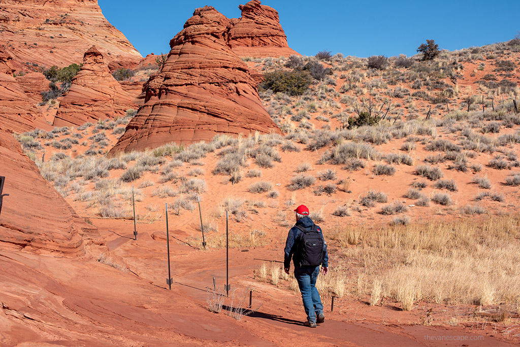 Chris Labanowski, co-founder of the Van Escape blog, is on the hiking trail to Coyote Butte South, he is wearing black backpack and hiking boots and red hat.