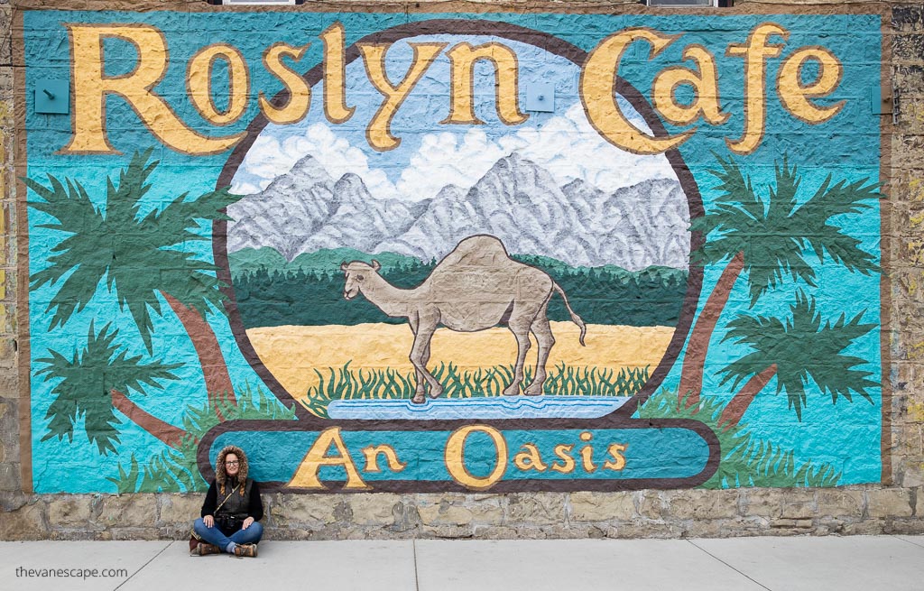 Agnes Stabinska, the author and co-founder of The Van Escape blog, is sitting in front of a mural wall; a camel is drawn on the wall, palm trees and mountains in the background, and the inscription: Roslyn Cafe and Oasis is a scenery for Northern Exposure series.