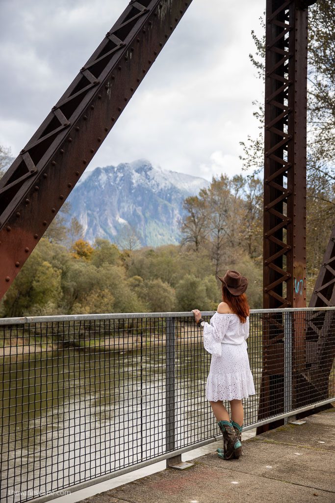 Agnes Stabinska, the author and co-owner of the Van Escape blog, is on the Ronette’s Bridge from Twin Peaks. She is looking att the river and mountains, she is wearing brown cowboy hat, brown cowboy boot and white dress. 