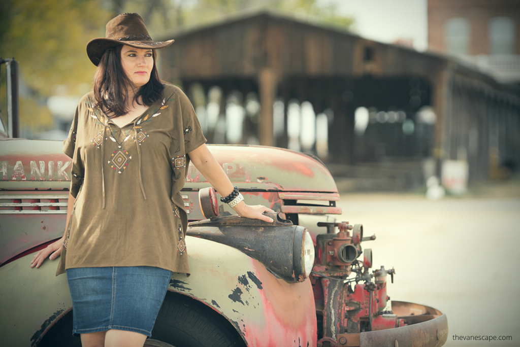 Agnes Stabinska, the author and co-owner of the Van Escape blog, wears cowboy hat in Shaniko Ghost Town.  She is next to old car and with old wooden buildlings in the backdrop.