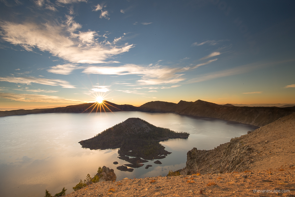 A stunning view of the sun rising behind the rock above the Crater Lake ridge. The rocks are in the warm yellow rays of the sun, in the middle of the lake is Wizard Island,