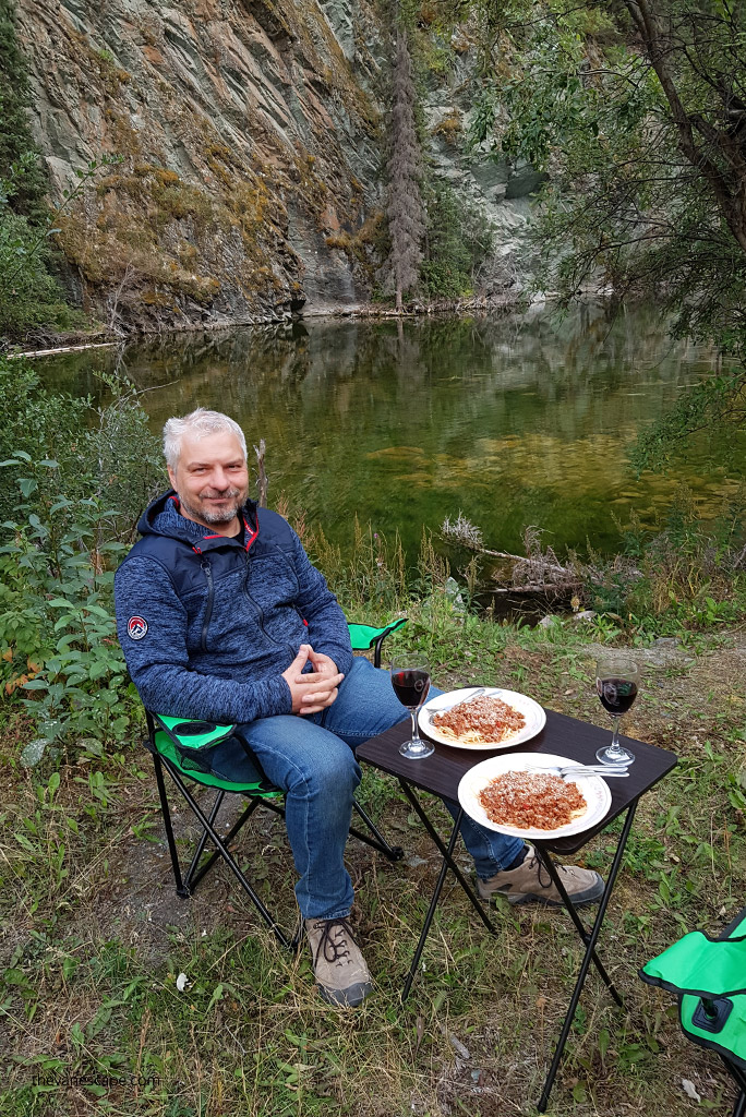 Chris Labanowski is sitting on the green camping chair next to black camping table with the river and mountain view. On the table are two camping wine glasses with red wine inside and two camping plates with spaghetthi on it.