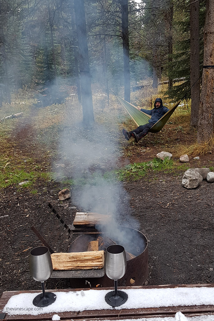 Chris Labanowski is in a green hammock between trees on the campfire. There is a table with camping wine glasses on it.