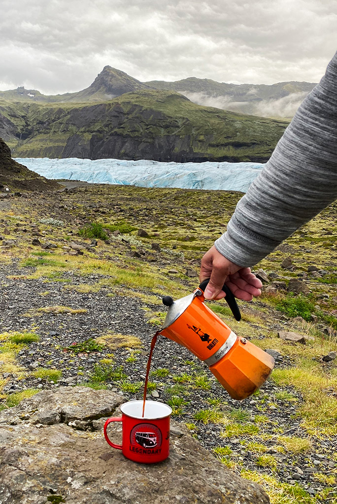 Hot black coffee is poured into a tin mug from an orange Bialetti coffee maker. A glacier and mountain view is in the background.