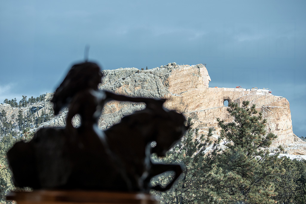 in the foreground, a bronze Crazy Horse figurine standing on a platform in the museum, and in the background, a huge scale in which work on the Crazy Horse sculpture is still ongoing.