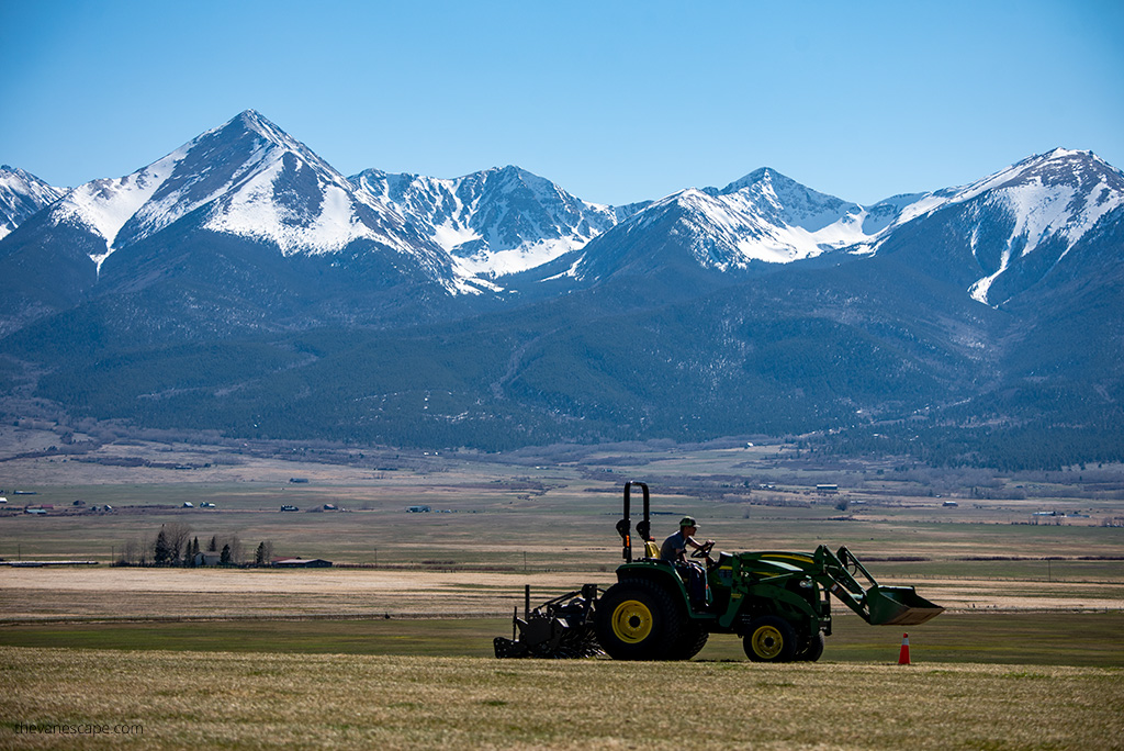 rulal view in Westcliffe: a man is driving a green tractor in the background there are majestic snow-capped mountain peaks