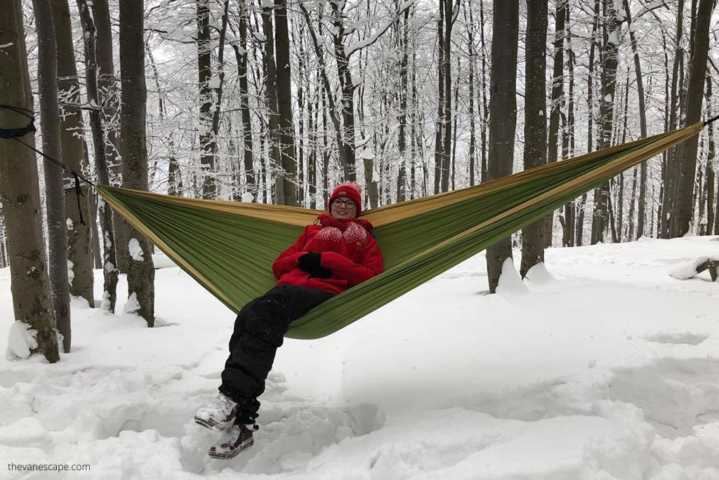 Agnes in winter clothes, warm red jacket and red hat in green  Backpacking Hammock hanging betweek trees in winter scenery.