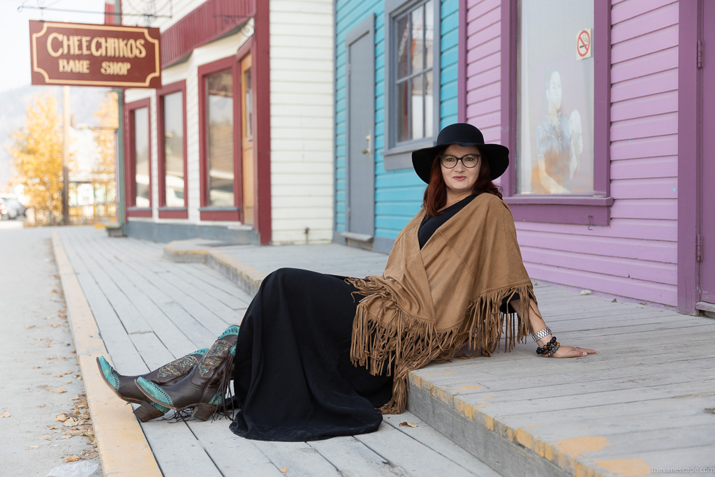 Agnes Stabinska, the author, in cowboy boots, long black dress, black hat, brown poncho, is sitting on the wooden  broadwalk in Dawson City.