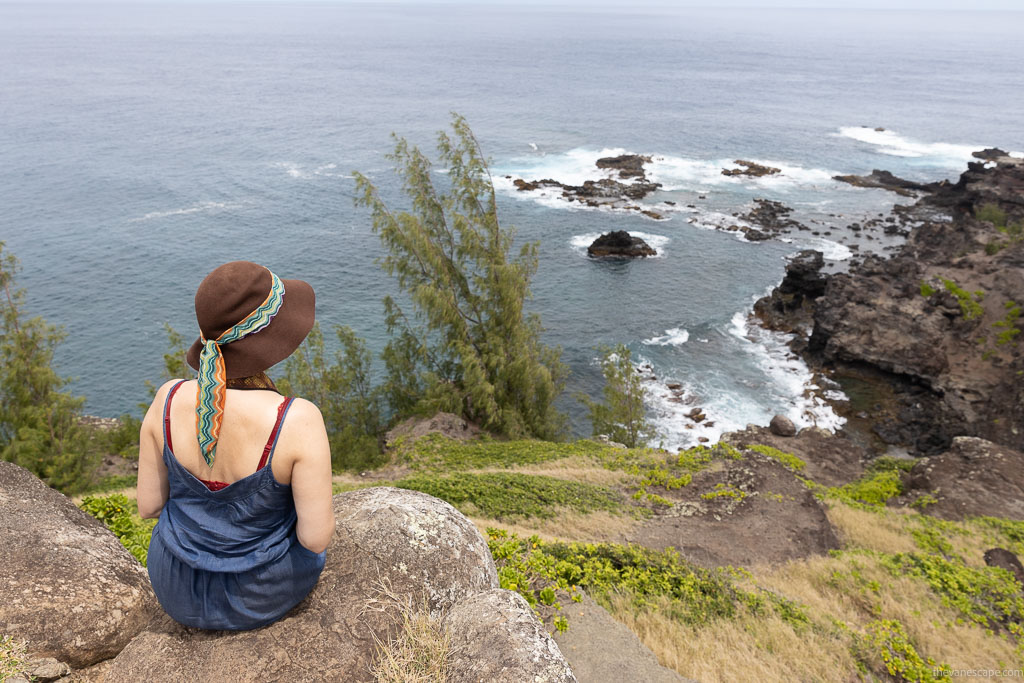 Agnes Stabinska, an author, sitting on the rock, wearing a blue dress and a brown hat and amiring sea view on Maui.  