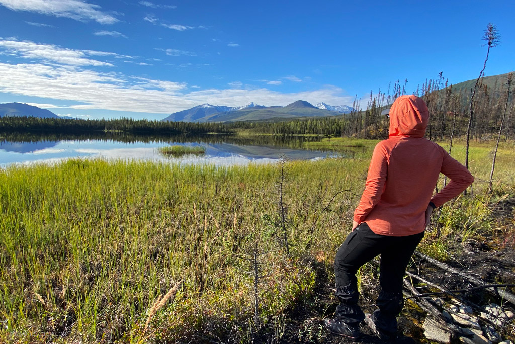 Agnes Stabinska, the author and co-founder of The Van Escape blog is standing on a trail in mountain, among grass with the lake and mountain view. She is wearing orange KUHL ENGINEERED Women's Hoody and black pants.