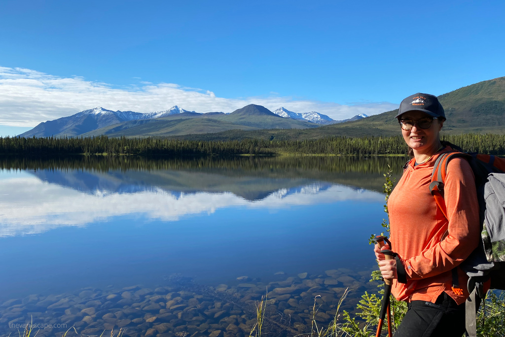 Agnes Stabinska, the authoor, in orange KUHL ENGINEERED Women's Hoody hiking in the mountains in Alaska. She is standing next to blue lake with mountains in the backdrop and mountains reflaction in the lake.