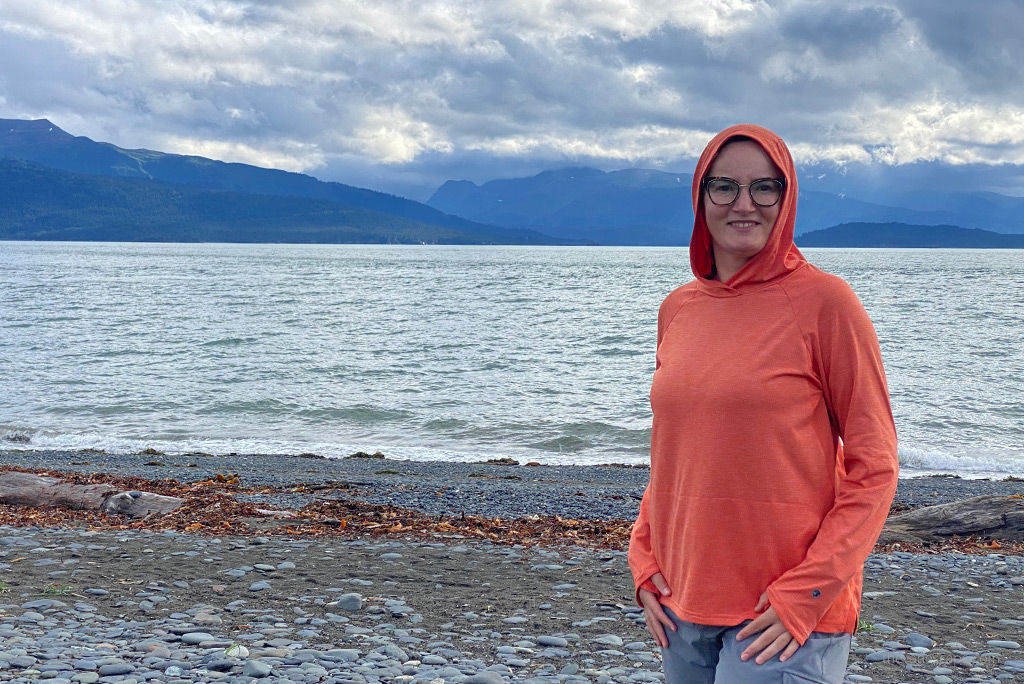 Agnes Stabinska in orange hoodie in Homer, Alaska with the sea and mountains in the backdrop.