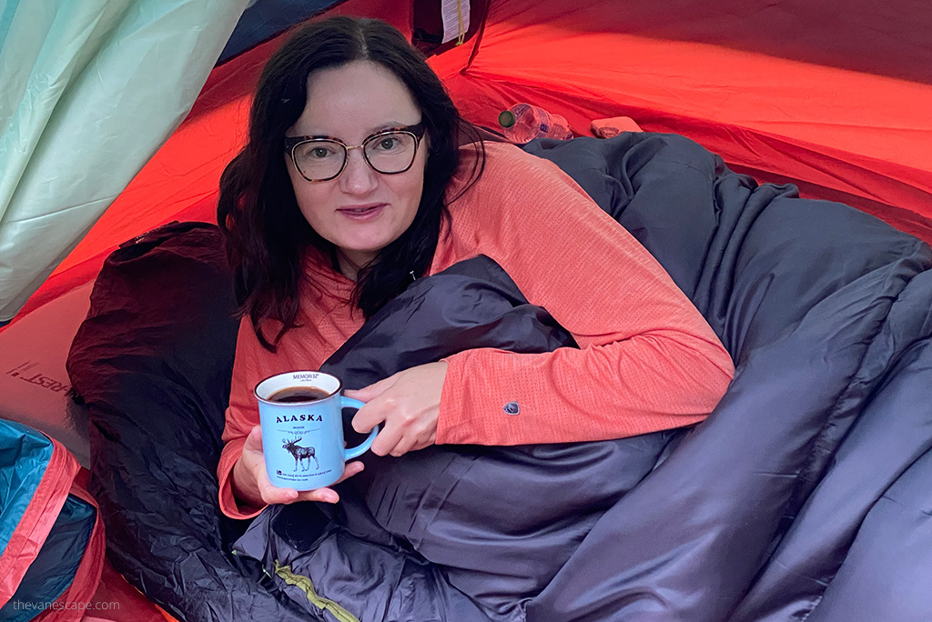 Agnes Stabinska, in orange KUHL ENGINEERED Women's Hoody is in a tent in Alaska in a sleeping bag and she is drinking coffee.