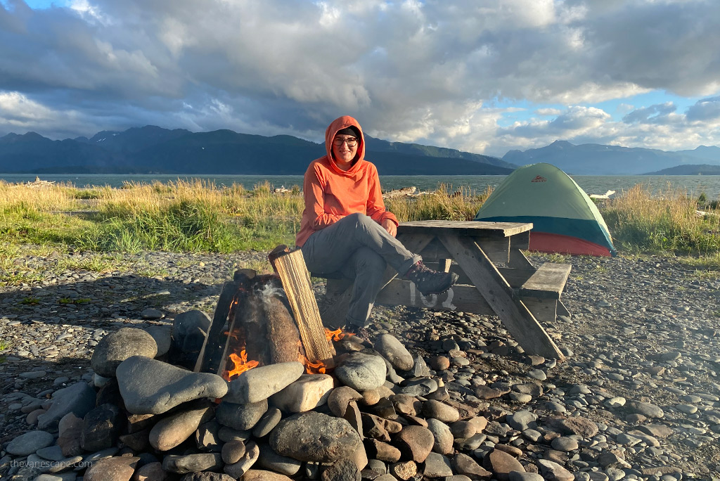 Agnes Stabinska, the author, is sitting in orange KUHL ENGINEERED Women's Hoody next to campfire with the tent and sea and mountains in the backdrop.