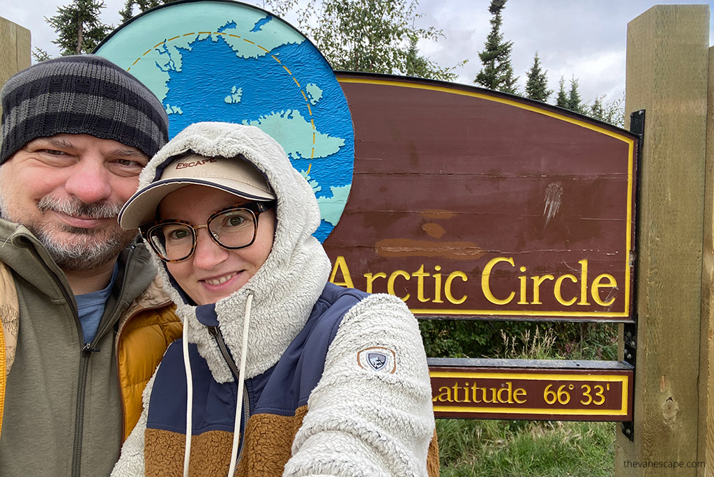 Agnes Stabinska with her partner Chris, owners of The Van Escape blog, are standing next to wooden sign of Arctic Circle in Alaska. Agnes wears hoody by Kuhl.