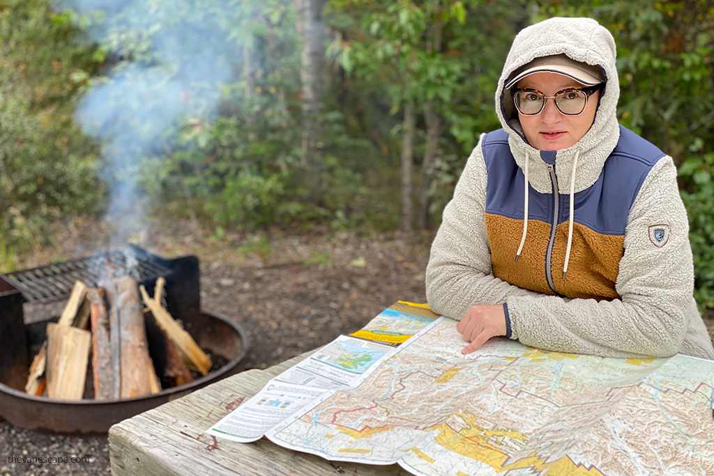 Agnes Stabinska, the author and co-owner of The Van Escape blog, is wearing KUHL PRISM Hoody and she is sitting next to campfire at the picnic table and she is looking at the hiking trail map.