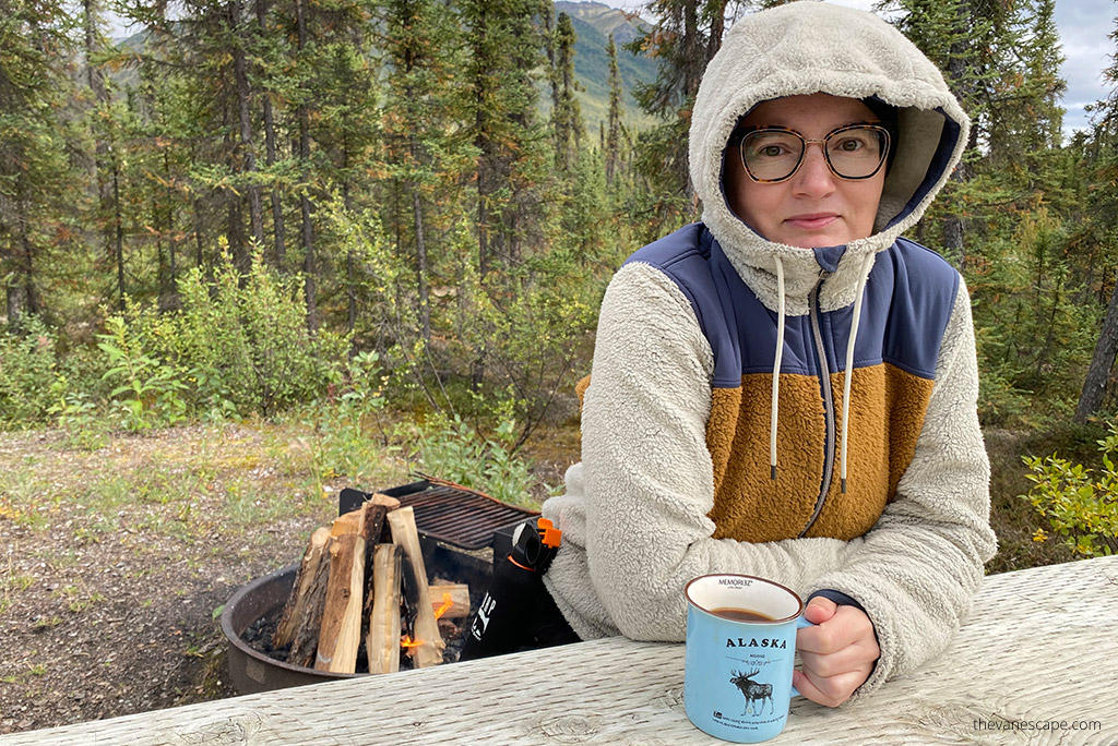 Agnes Stabinska, the author in Women’s KUHL PRISM Hoody is sitting next to campfire with the mountain view and she is drinking coffee in a mug with inscription: Alaska.