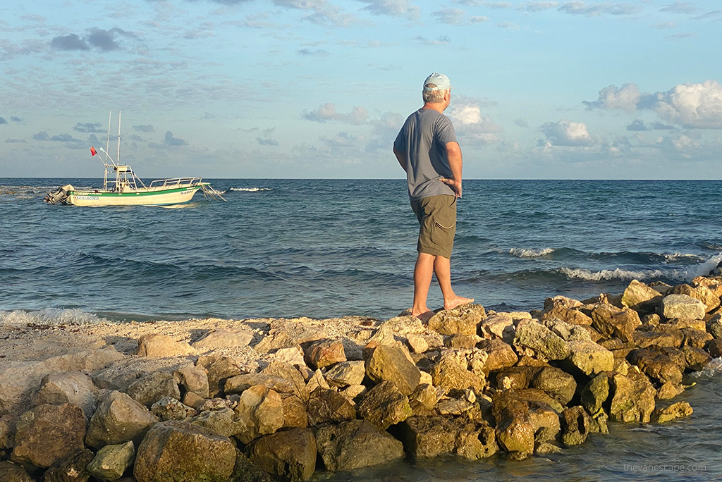 Chris Labanowski, co-founder of The Van Escape blog, is waring KUHL AMBUSH Cargo Shorts. He is at the rocks on the beach with the sea view and boat in the backdrop.