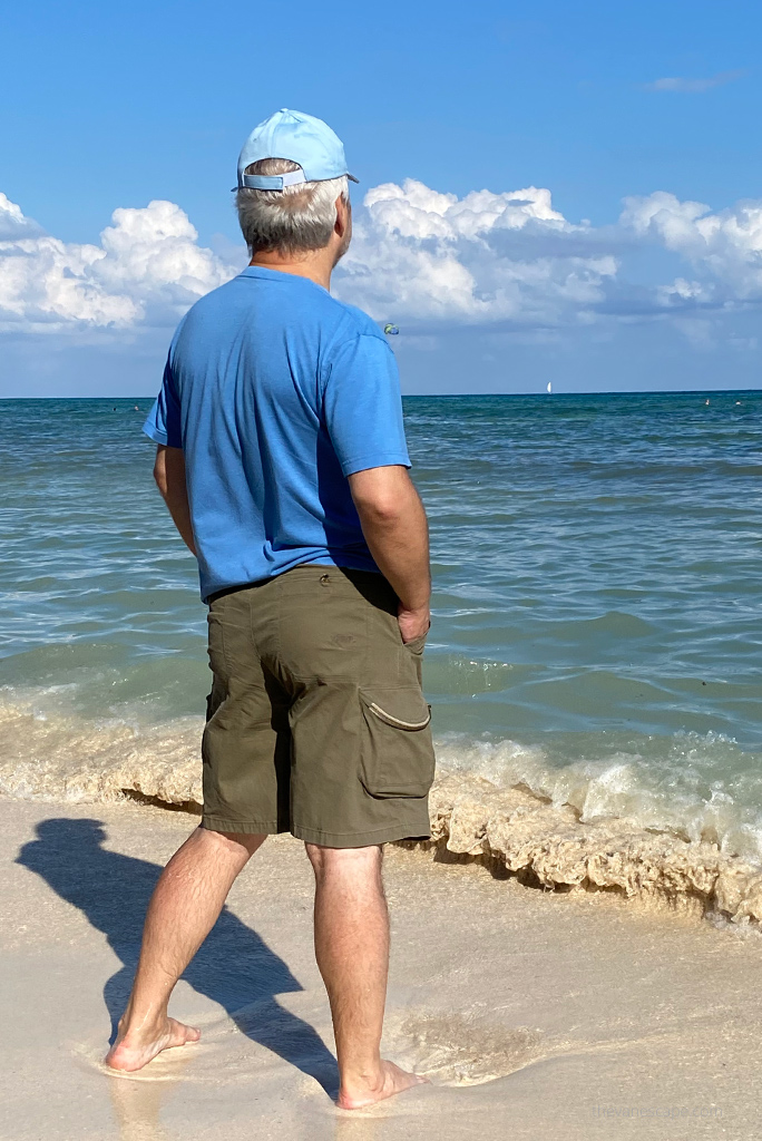 Chris Labanowski, in a blue T-shirt, blue baseball cap, and olive AMBUSH Cargo shorts, walks on a sandy beach on the Caribbean Sea in Mexico.