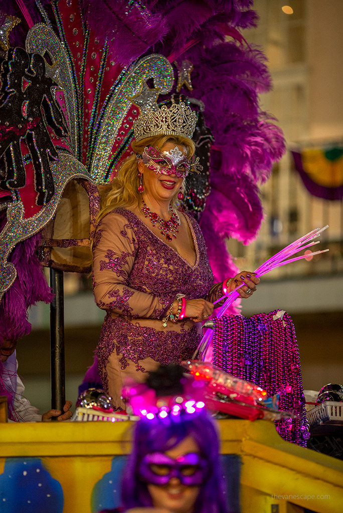 a woman in a large purple headdress and a mask waving to the crowd