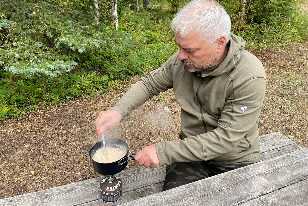 Chris Labanowski, co-founder of the Van Escape blog, is sitting on the picnic table, at the campsite and he is making a dinner at the camping stove.