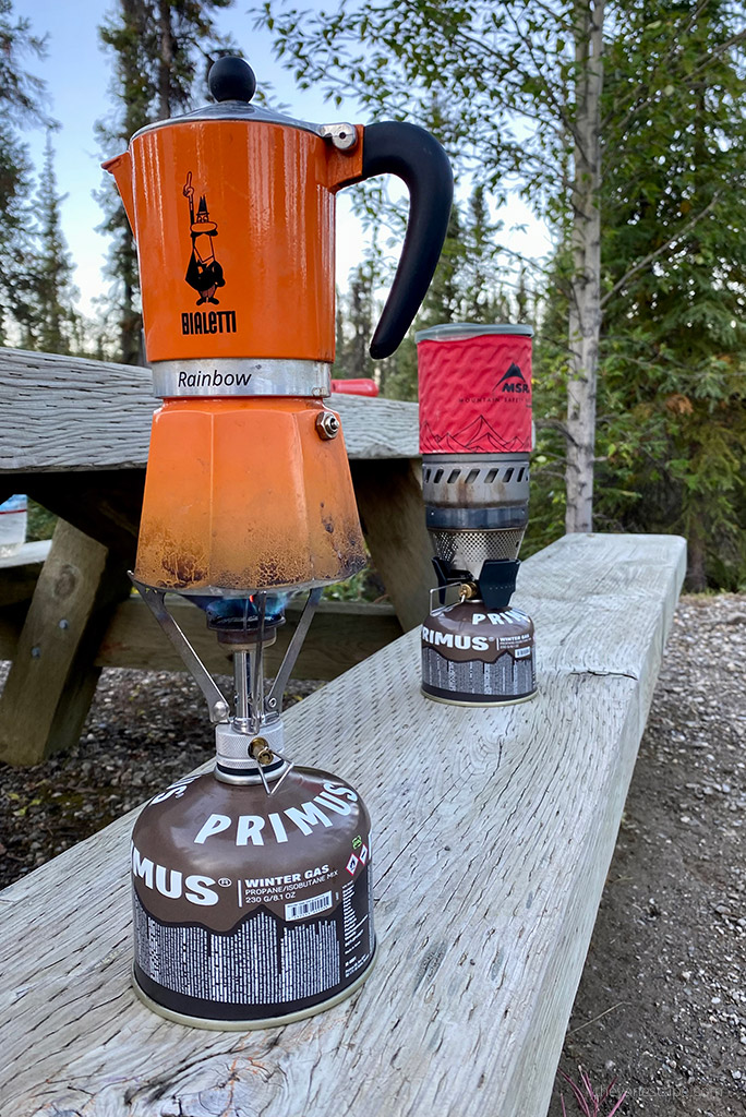 The Orange Bialetti cafe maker is on a fuel gas stove, and the red MSR camping stove is on a fuel gas stove on a wooden bench at the campsite.