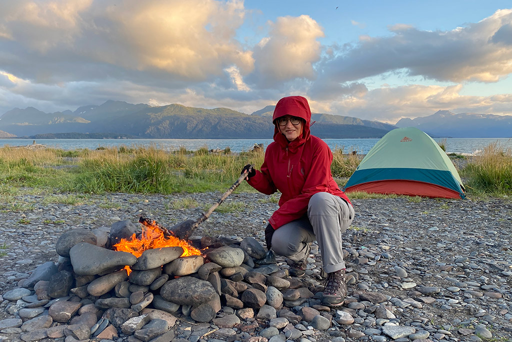 KUHL FREEFLEX ROLL-UP Pants Review: Agnieszka Stabinska, the author, in gray pants by Kuhl, is lighting a fire at a campsite by the sea. At the back is a tent and the mountains, it's Alaska.