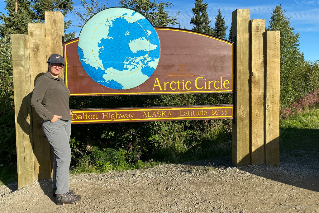 Agnes Stabinska, the author, is wearing Kuhl pants and she is standing at the wooden Arctic Circle sign in Alaska.