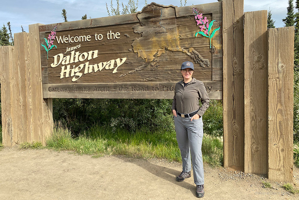 Agnes Stabinska, the author and co-founder of the Van Escape blog is standing next to wooden sign of the Dalton Highway in Alaska. She is wearing gray KUHL FREEFLEX ROLL-UP Pants. 