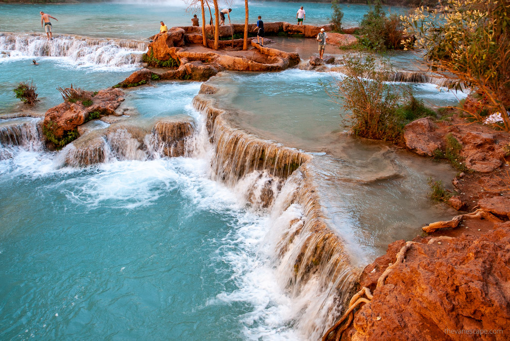 People wading and playing in the shallow blue water of Havasu Falls among the green trees and rust-colored canyon walls.
