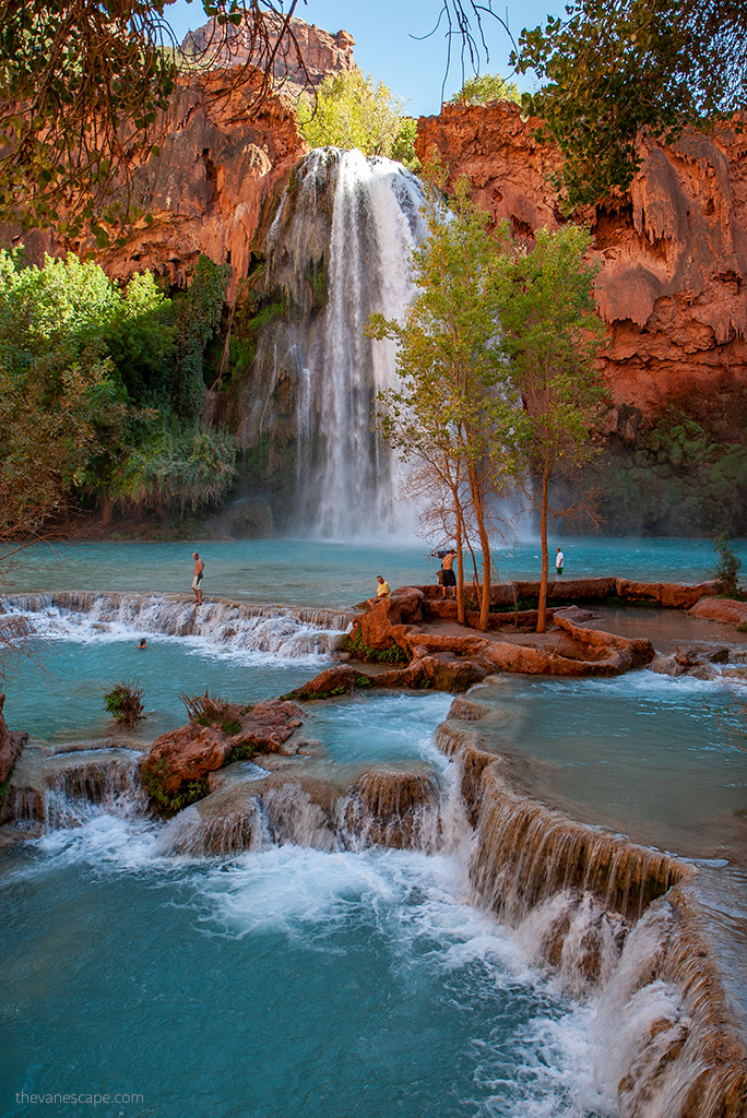 stunning blue water of Havasu Falls flowing down high orange rocks, greenery all around and people playing in the water.