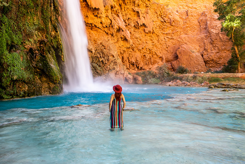 Agnes Stabinska, the author, stands in a colorful striped dress, immersed up to her calves in the blue water of Havasu Falls, and admires the cascading water from orange rocks.