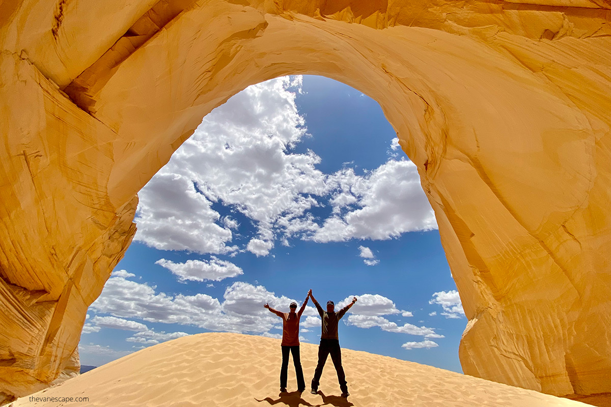 Agnes Stabinska, the author with her partner Chris Labanowski in Great Chamber near Kanab. Theay are standing on a sandy dune, the walls are yellow, the sky is blue. 