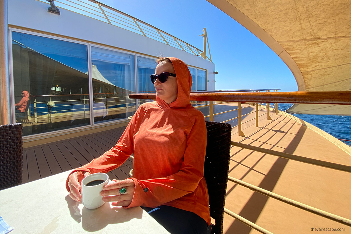 Agnes drinking coffee at ship during cruise, wearing orange Kuhl sun protection t-shirt with a hood and sunglasses